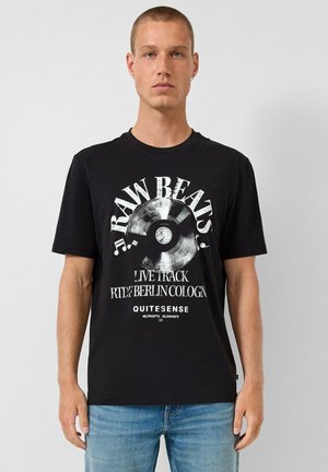 Jeune homme portant un t-shirt noir avec un disque vinyle blanc et le texte « RAW BEATS, LIVE TRACK, BERLIN, COLOGNE, QUITESENSE », associé à un jean bleu.