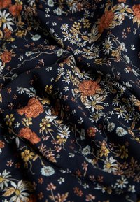 Blomstret stof har en mørk navy baggrund med intrikate mønstre af orange, gule og hvide blomster. Tekstureret, blød kvalitet.