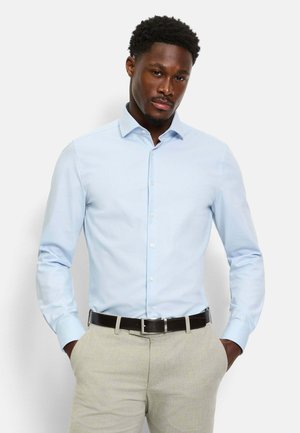 Homme portant une chemise à manches longues bleu clair boutonnée, rentrée dans un pantalon beige avec une ceinture noire, debout les mains dans les poches.