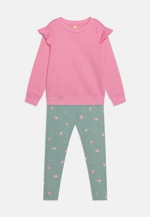 Roza majica z dolgimi rokavi z naborki na ramenih v kombinaciji z zelenimi legicami z majhnim rožnatim cvetličnim vzorcem.