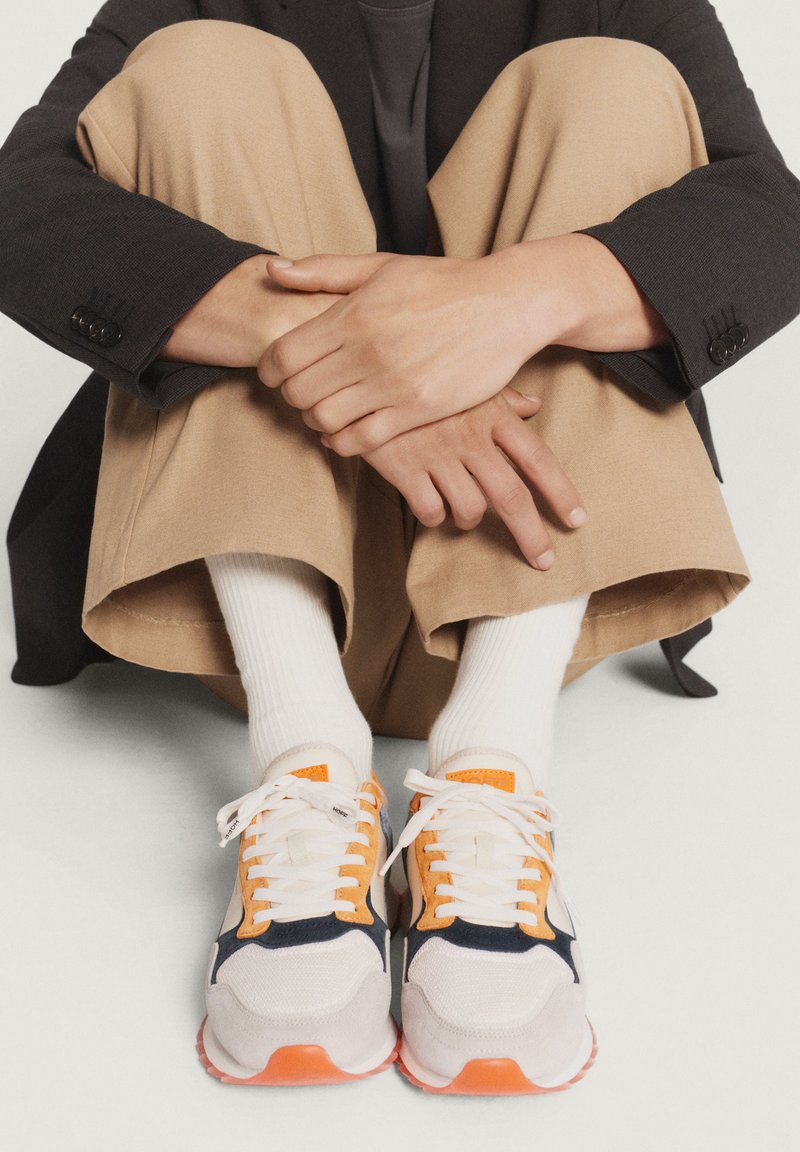 Sneakers eleganti con una parte superiore in rete bianca, dettagli blu navy e arancioni, suole testurizzate e lacci piatti, posizionati in una posa seduta.