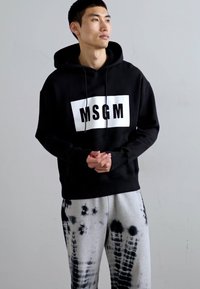 Schwarzer Hoodie mit weißem Logo auf der Vorderseite, Kapuze mit Zugband und gerippten Bündchen. Kombiniert mit grauen Tie-Dye-Sweatpants mit schwarzen Mustern.