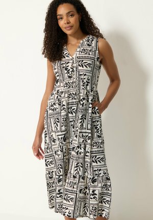 Mujer con cabello rizado que lleva un vestido midi sin mangas con estampado en blanco y negro y bolsillos, de pie frente a un fondo claro.