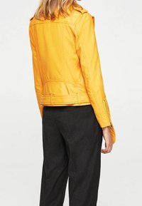 Žlutá kožená moto bunda s cropped designem, přiléhavým střihem, ramenními epauletami a zipovými manžetami. Nosič má na sobě černé kalhoty.