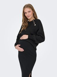 Nėščia moteris su ilgais plaukais, dėvinti juodą megztinį ir juodą, ribotą sijoną, švelniai glostanti pilvuką ir švelniai šypsosi.