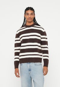 Pull rayé marron et blanc avec un col rond, des poignets et un ourlet côtelés. Porté avec un jean bleu clair, en matériau tricoté texturé.