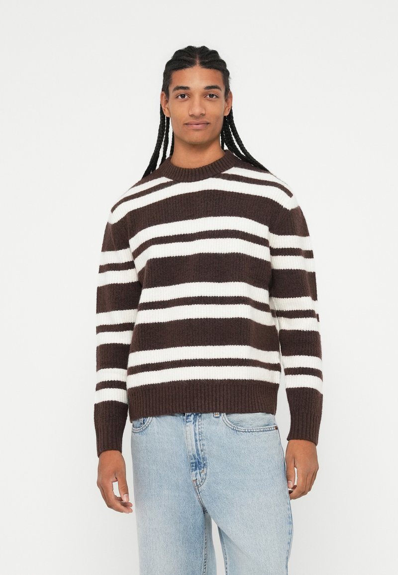 Pull rayé marron et blanc avec un col rond, des poignets et un ourlet côtelés. Porté avec un jean bleu clair, en matériau tricoté texturé.