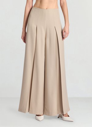 Pantaloni a gamba larga in tessuto beige chiaro, con pieghe frontali e una texture liscia. Abbinati a tacchi bianchi a punta.