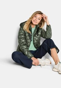 Grüne, gesteppten Puffermantel mit Kapuze, der einen vollständigen Frontreißverschluss und schwarze Beschläge aufweist. Getragen über einem hellgrünen Pullover und dunklen Jeans.