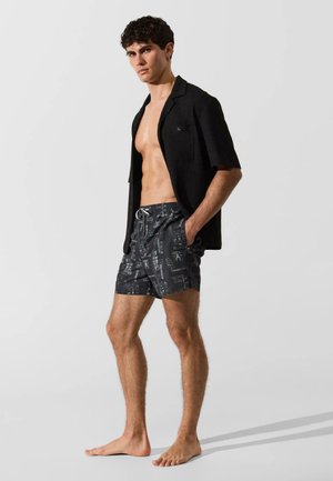 Mladý muž stojí bosý, má na sebe otvorenú čiernu krátku košeľu a čierne vzorované plavky s rukami v vreckách.