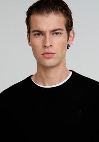 Suéter negro de punto con cuello redondo y cuello interior blanco. Presenta un pequeño logo bordado en el pecho. Textura suave.