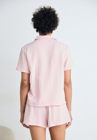 Pajama set s kratkim rukavima i ovratnikom u nježnoj ružičastoj boji, izrađen od teksturiranog materijala. Šortsevi imaju elastičnu traku u struku za udobnost.