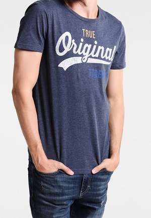 Homme portant un T-shirt bleu marine avec le texte "True Original" et un jean bleu, debout les mains dans les poches sur un fond blanc.