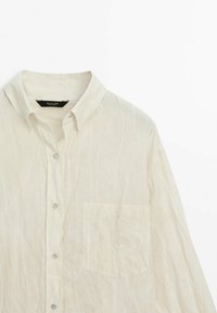 Chemise blanche à manches longues avec col et poche poitrine, fabriquée en tissu léger légèrement froissé.