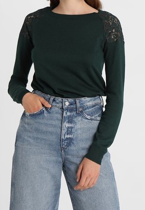 Nainen pukeutuneena tummanvihreään pitkähihamaiseen neuleeseen, jossa on pitsikoristeiset olkapäät, ja vaaleansiniset korkeavyötäröiset farkut.