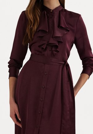 Kobieta ubrana w bordową, długorękawową sukienkę na guziki z falbankami z przodu i paskiem wiązanym w talii, stojąca z jedną ręką na biodrze.