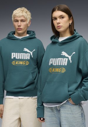 Dos modelos que llevan sudaderas con capucha Puma King color verde azulado, uno con pantalones claros y cabello rubio, y el otro con jeans y cabello castaño, parados uno al lado del otro.