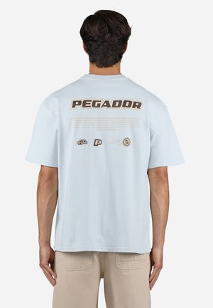 Helles blaues Baumwoll-T-Shirt mit kurzen Ärmeln, das auf der Rückseite den Text "PEGADOR" in fetten, dunklen Buchstaben sowie zusätzliche grafische Details zeigt.
