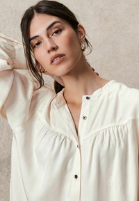 Lichtbeige blouse met wijd uitlopende mouwen, geplooide halslijn en knoopsluiting met zilveren accenten en subtiele plooidetails.