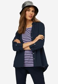 Camisa de botones de manga larga en color marino sobre una camiseta de rayas moradas, combinada con pantalones oscuros y un sombrero de cubo plateado brillante.