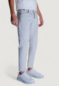 Jeans de corte ajustado en gris claro, confeccionados en algodón, con cierre de botón y cremallera, diseño de cinco bolsillos y detalles de costura sutiles, combinados con zapatillas blancas.