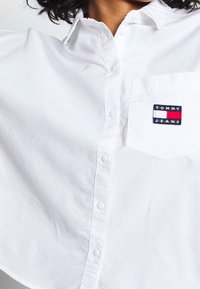 Vit skjorta med knappar och bröstficka. Har "TOMMY JEANS"-logotyp i svart, röd och vit. Slät tyg med en klassisk kragsdesign.