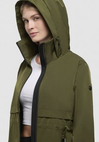 Olivovo zelená bunda na zips s kapucňou, s čiernymi akcentmi, vysokým golierom a dvoma prednými vreckami. Hladký materiál a moderný dizajn.