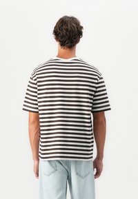 Mann med kort krøllete hår står med ryggen til iført en brun og hvitstripete kortermet skjorte og lyseblå jeans.