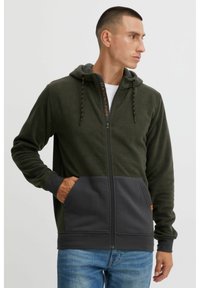 Sudadera con cremallera de forro polar verde, con una sección inferior gris, cordones en contraste y bolsillos frontales. Textura suave, diseño informal.