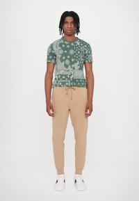 Groene gemêleerde short-sleeve t-shirt met witte ontwerpen, gecombineerd met beige joggingbroek met trekkoord en witte sneakers.