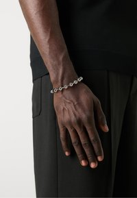 Silver metal bracelet with spherical beads, worn on a wrist, alongside a black shirt and pants, highlighting a smooth, reflective surface.