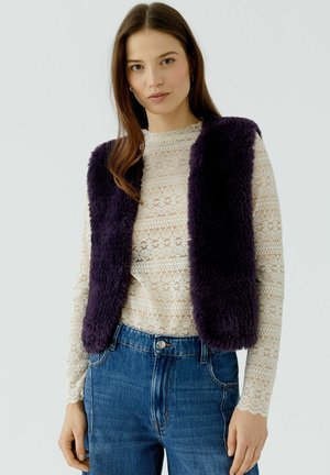 Femme portant un gilet en fausse fourrure violet foncé sur un haut blanc en dentelle à manches longues et un jean bleu, debout devant un fond clair uni.