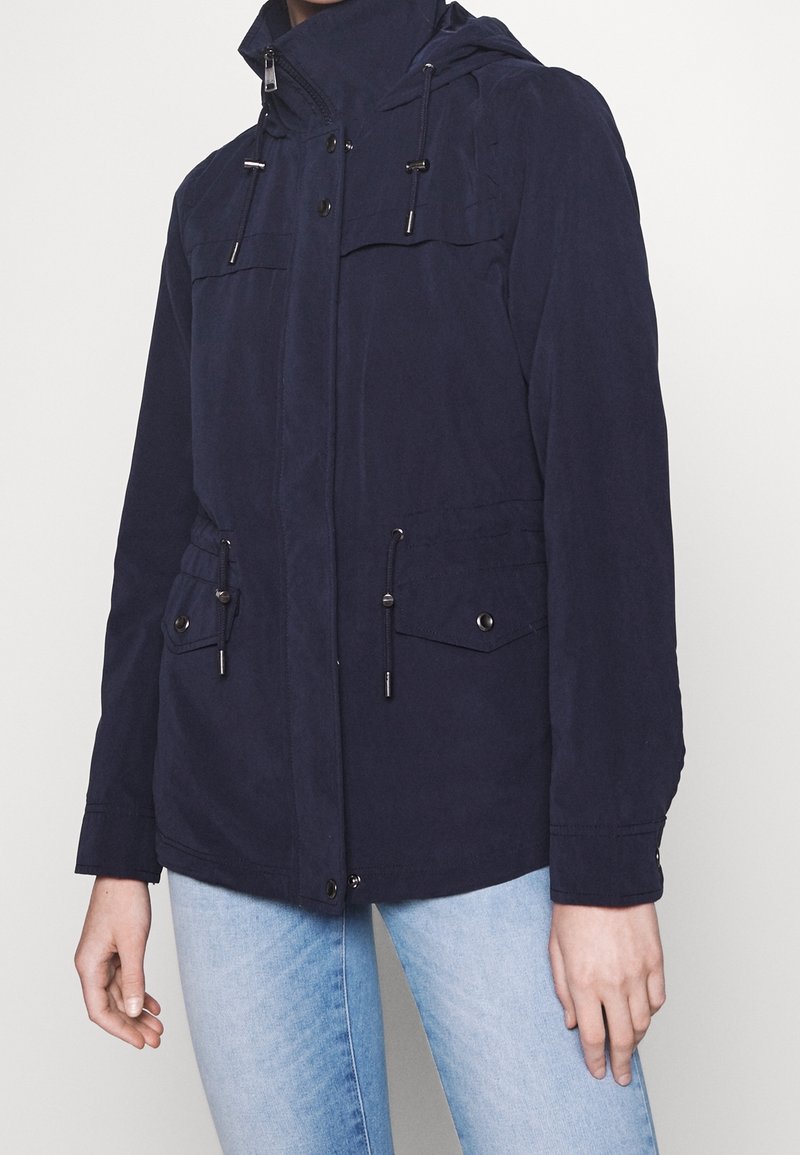Veste à capuche bleu marine avec poches avant, taille ajustable par cordon et fermeture éclair. Confectionnée en tissu léger à la texture lisse.