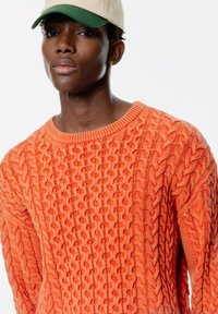 Orange stickad tröja med ett strukturerat kabelmönster. Ribbad halsringning och ärmslut, parat med en beige keps med en grön skärm.