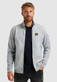 Grijze gewatteerde ritsjack met een staande kraag, voorzien van een logo-patch op de borst, zijzakken en geribde manchetten. Draagt over een donker T-shirt.
