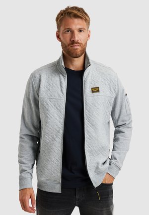 Harmaa quiltattu vetoketjullinen takki, jossa on noussut kaulus, logo-patch rinnassa, sivutaskut ja ribbikuvioidut hihansuut. Päällä tumma t-paita.