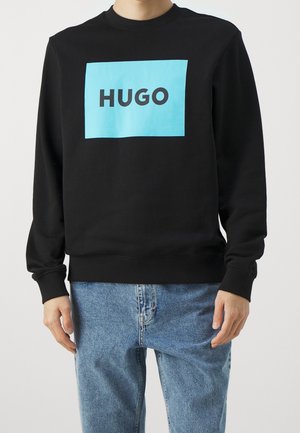 Mężczyzna w czarnej bluzie z dużym niebieskim kwadratem i napisem "HUGO" na piersi, w połączeniu z jasnoniebieskimi dżinsami.