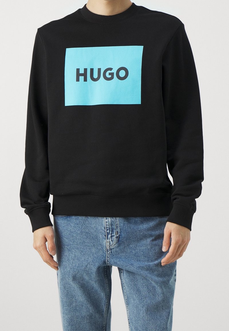 Homme portant un sweat-shirt noir avec un grand carré bleu et le texte "HUGO" sur la poitrine, associé à un jean bleu clair.