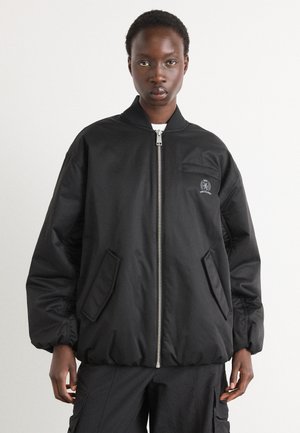 Chaqueta bomber negra con cierre frontal, cuello acanalado y bolsillos laterales. Presenta un logotipo sutil en el pecho y una textura lisa y mate.
