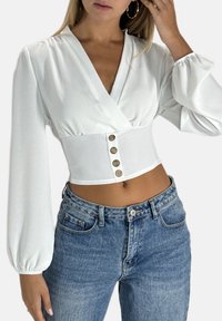White long-sleeve cropped blouse with a deep V-neck and five brown buttons at the waist. Worn with high-waisted light-wash jeans.