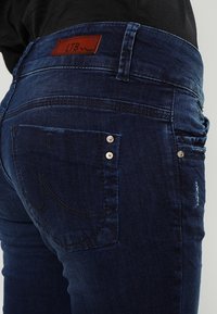 Jeans in denim blu scuro con vita media, caratterizzati da un'etichetta in pelle sul retro, due tasche con bottoni in metallo e leggere abrasioni.
