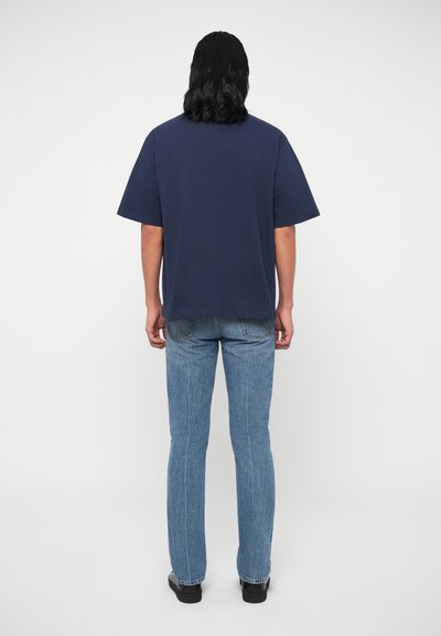 Námořnická modrá oversize bavlněná tričko s krátkými rukávy, kombinované s světlými modrými džíny, pohled zezadu na neutrálním pozadí.