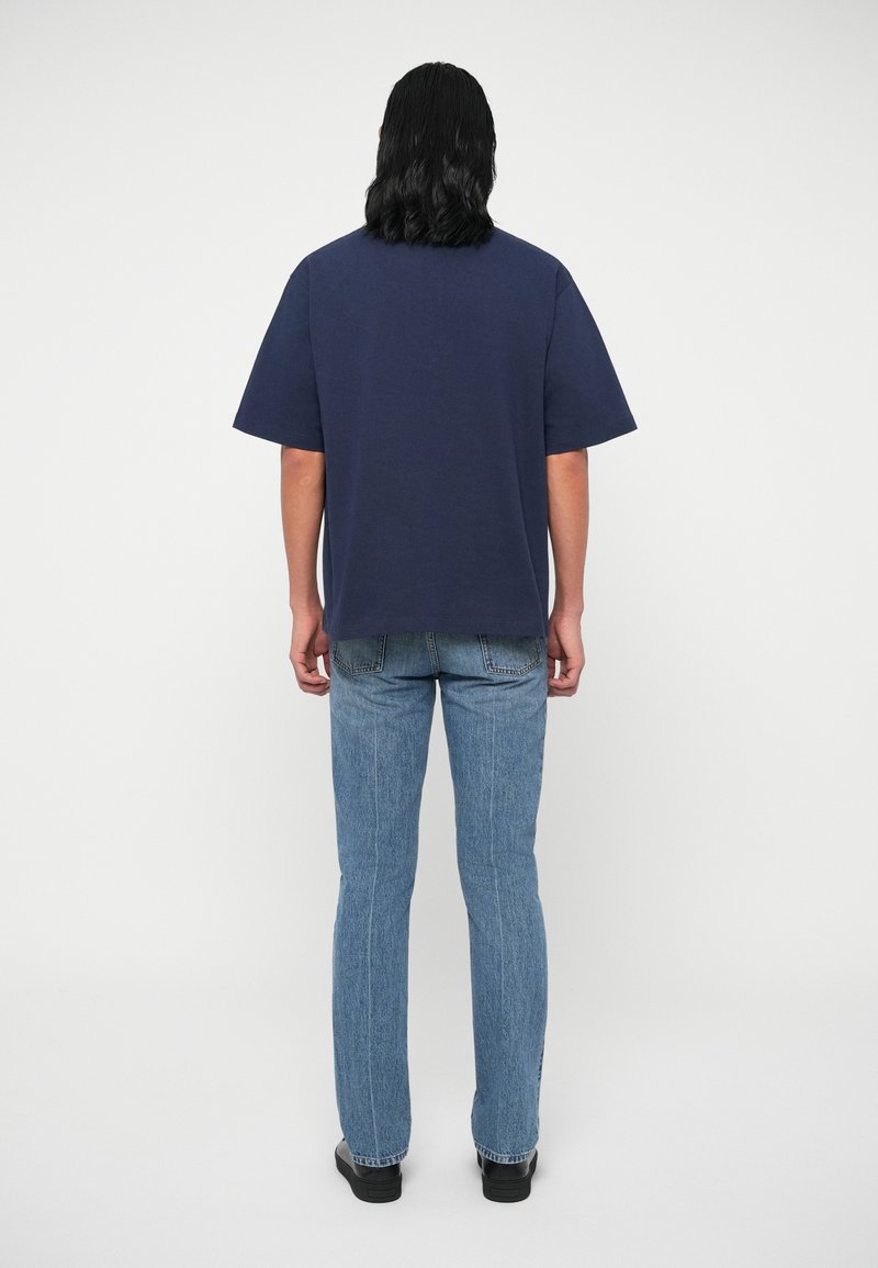 Dunkelblaues, oversized Baumwoll-T-Shirt mit kurzen Ärmeln, kombiniert mit hellblauen Jeans, von hinten auf einem neutralen Hintergrund betrachtet.
