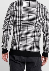 Schwarz-weißer karierter Pullover mit unterschiedlichen Mustern, gestrickter Textur, langen Ärmeln und einem schwarzen gerippten Bündchen und Saum.