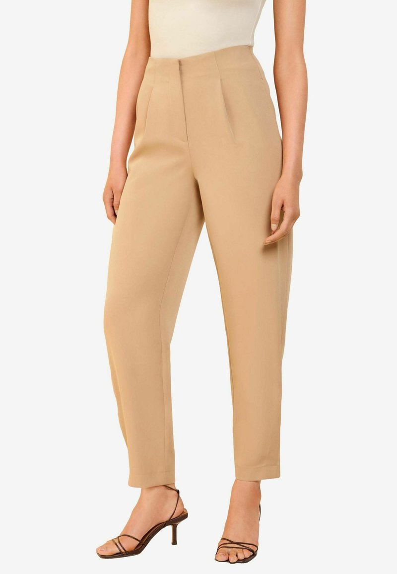 Beige high-waisted trousers with pleats, straight cut, and tapered leg. Smooth fabric with a matte finish. Worn with strappy high-heeled sandals.