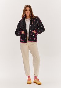 Čierna prešívaná bunda s farebnou kvetinovou výšivkou, ružovým lemom, zipsom a bočnými vreckami. V kombinácii so smotanovými nohavicami a žltými teniskami.