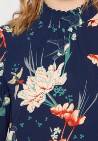 Marinblå blommig klänning med volangkrage, prydd med stora vita och orange blommor, gröna blad och ett mjukt, texturerat tyg.