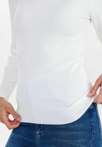 Weißes geripptes Langarmshirt, taillierte Passform mit abgerundetem Saum, getragen über blauen Jeans. Die Textur erscheint weich und leicht dehnbar.