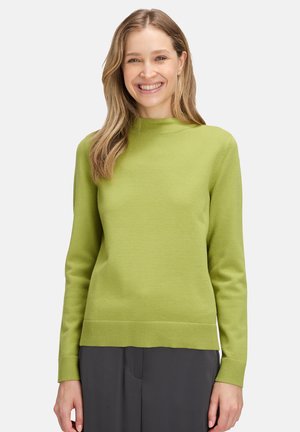 Lachende vrouw met lang blond haar draagt een lichtgroene lange mouwtrui en donkergrijze broek voor een effen witte achtergrond.