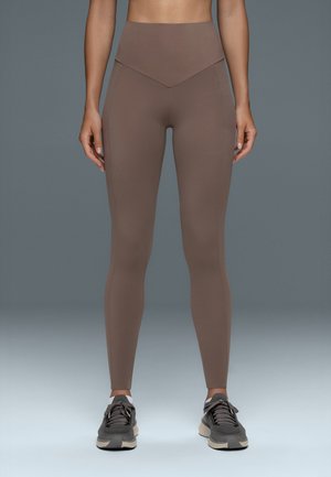 Femme portant des leggings marron taille haute et des chaussures de sport grises, debout devant un fond gris uni.